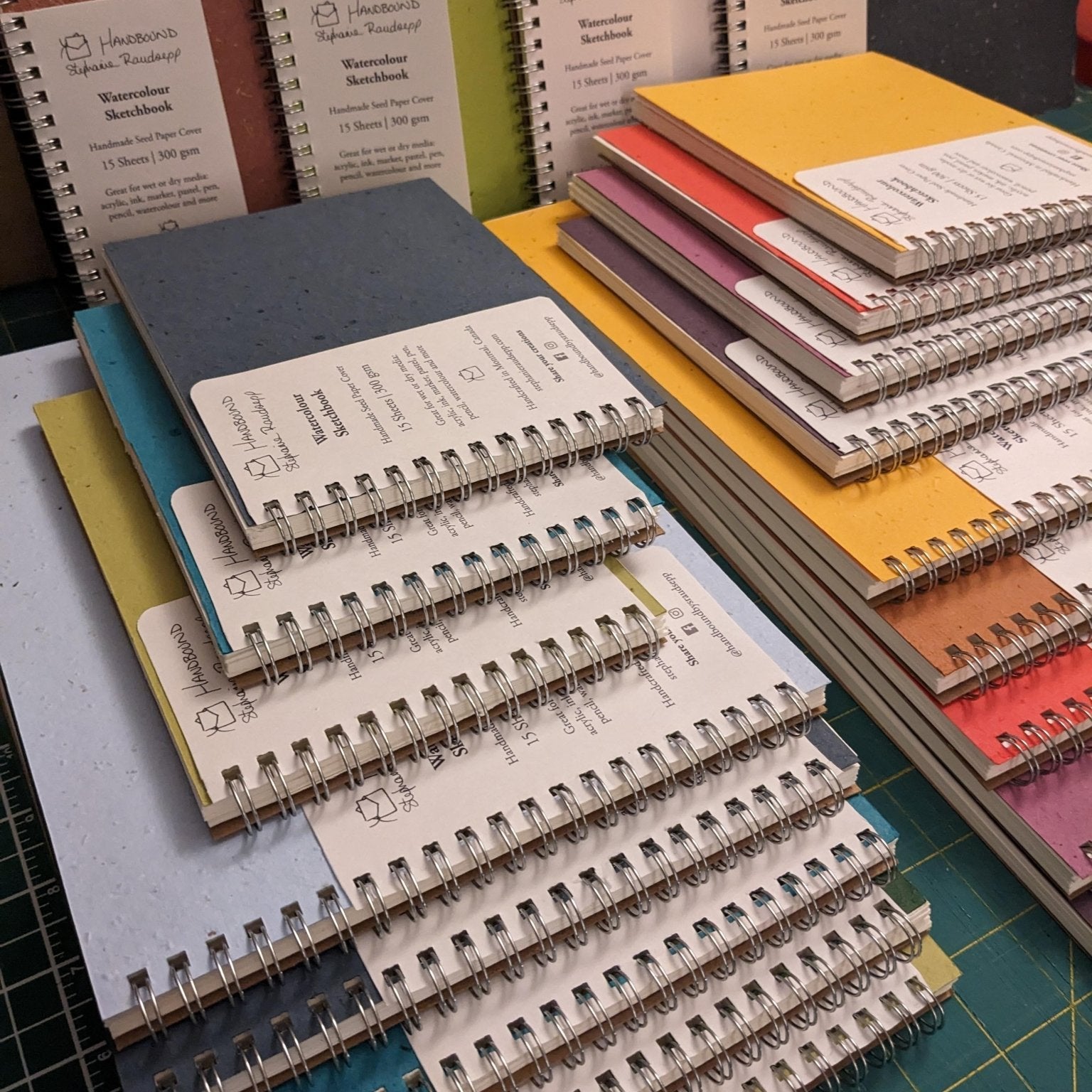 Stacks of colorful notebooks on a table with a cutting mat underneath. Large Plantable Watercolour Sketchbook by Creative Artifacts HQ
