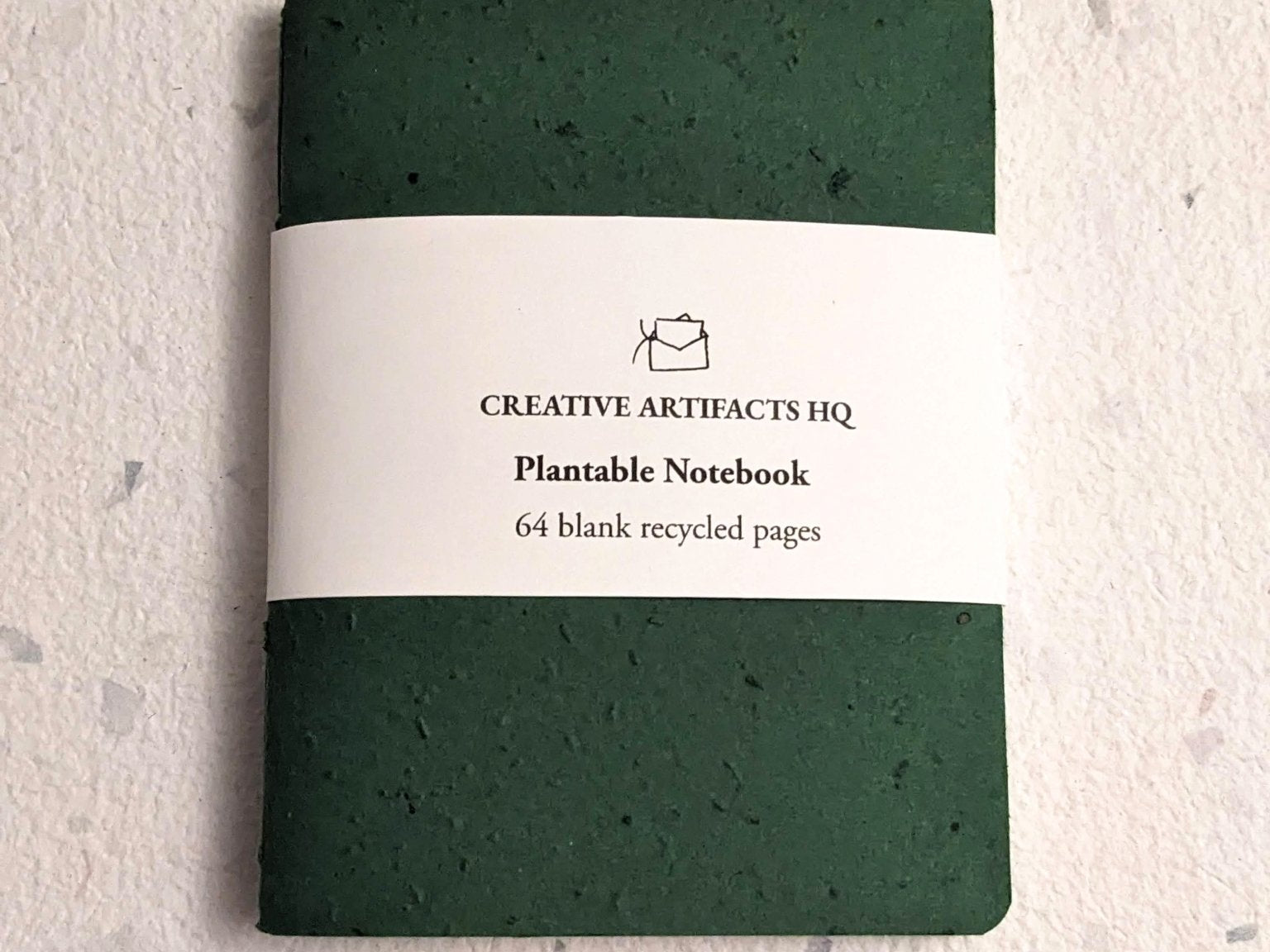 Green plantable notebook with a visible brand label on a textured hand stitched plantable paper notebook with string closure on a light grey background Creative Artifacts HQ
