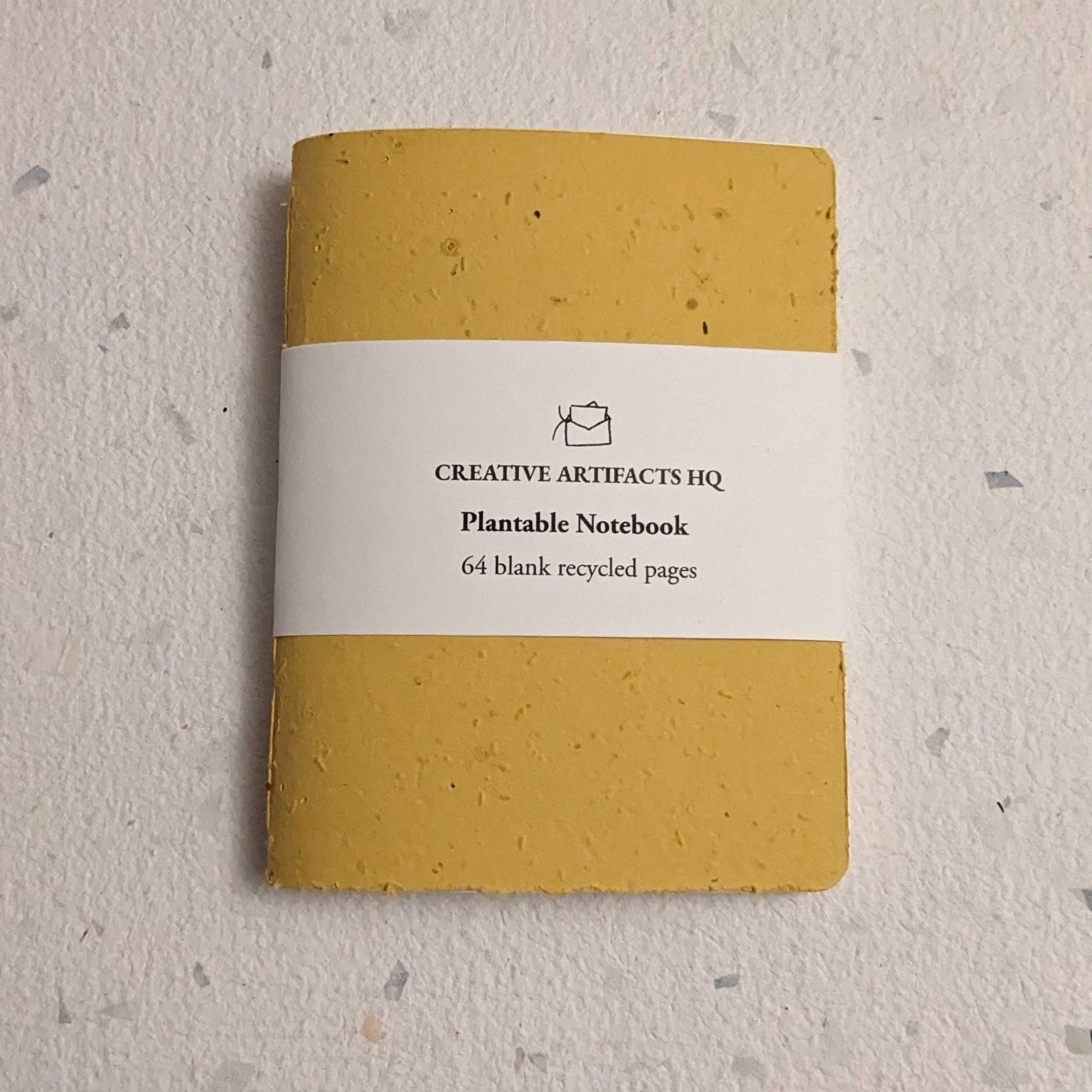 Yellow notebook with a white label on a hand stitched plantable paper notebook on a light grey background Creative Artifacts HQ