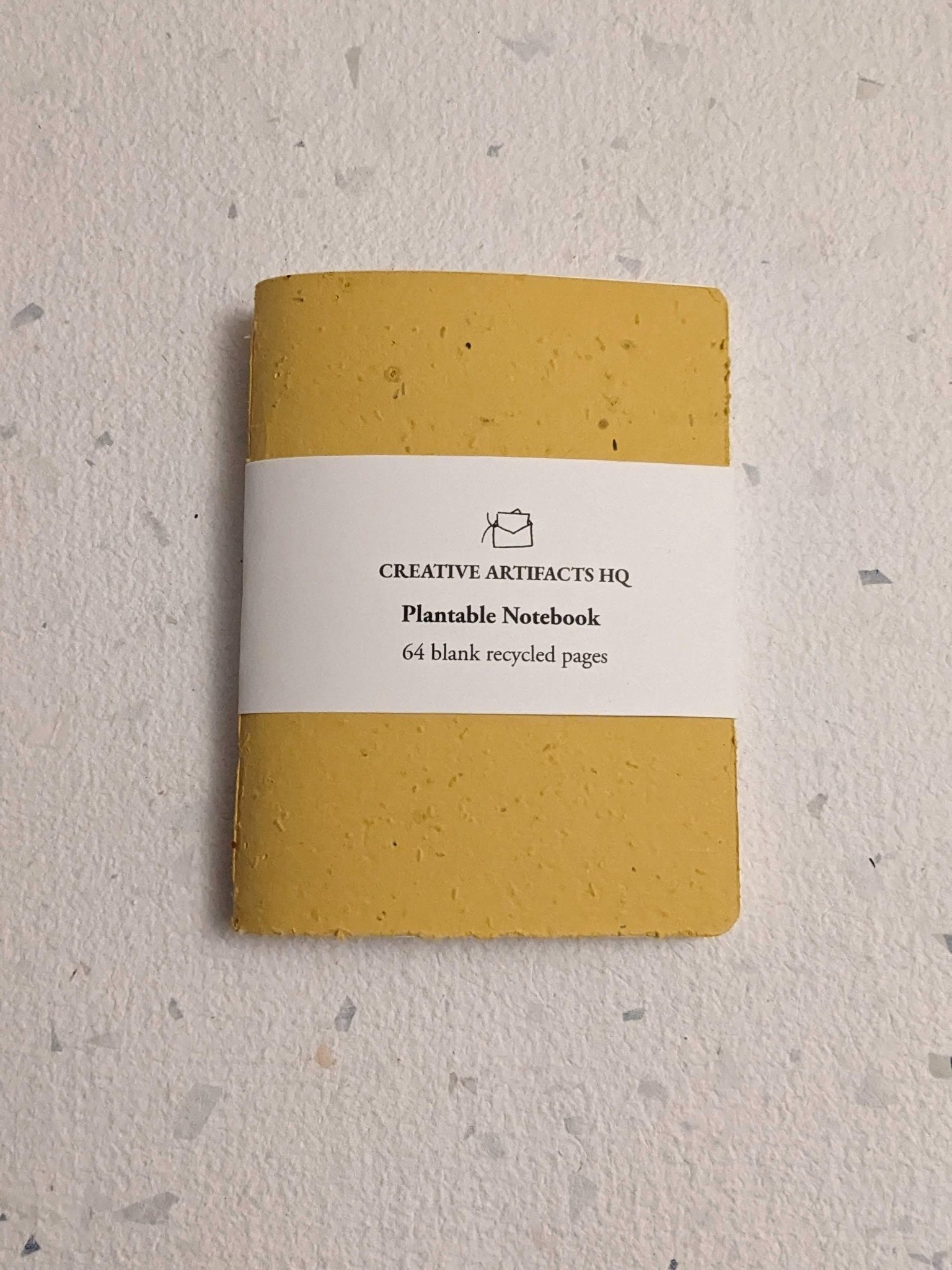 Yellow notebook with a white label on a hand stitched plantable paper notebook on a light grey background Creative Artifacts HQ