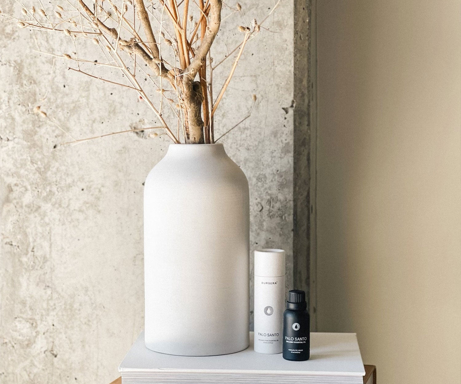 White vase with dried branches on a wooden surface against a textured wall.