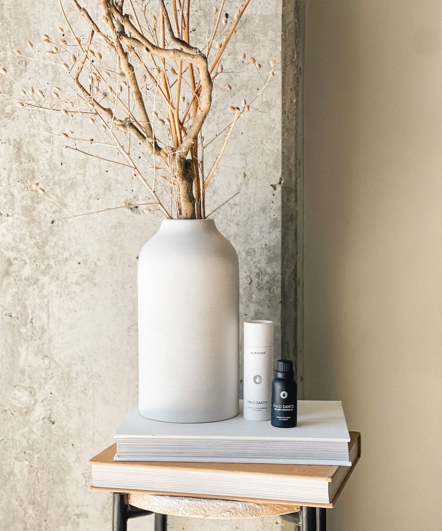 White vase with dried branches on a wooden surface against a textured wall.