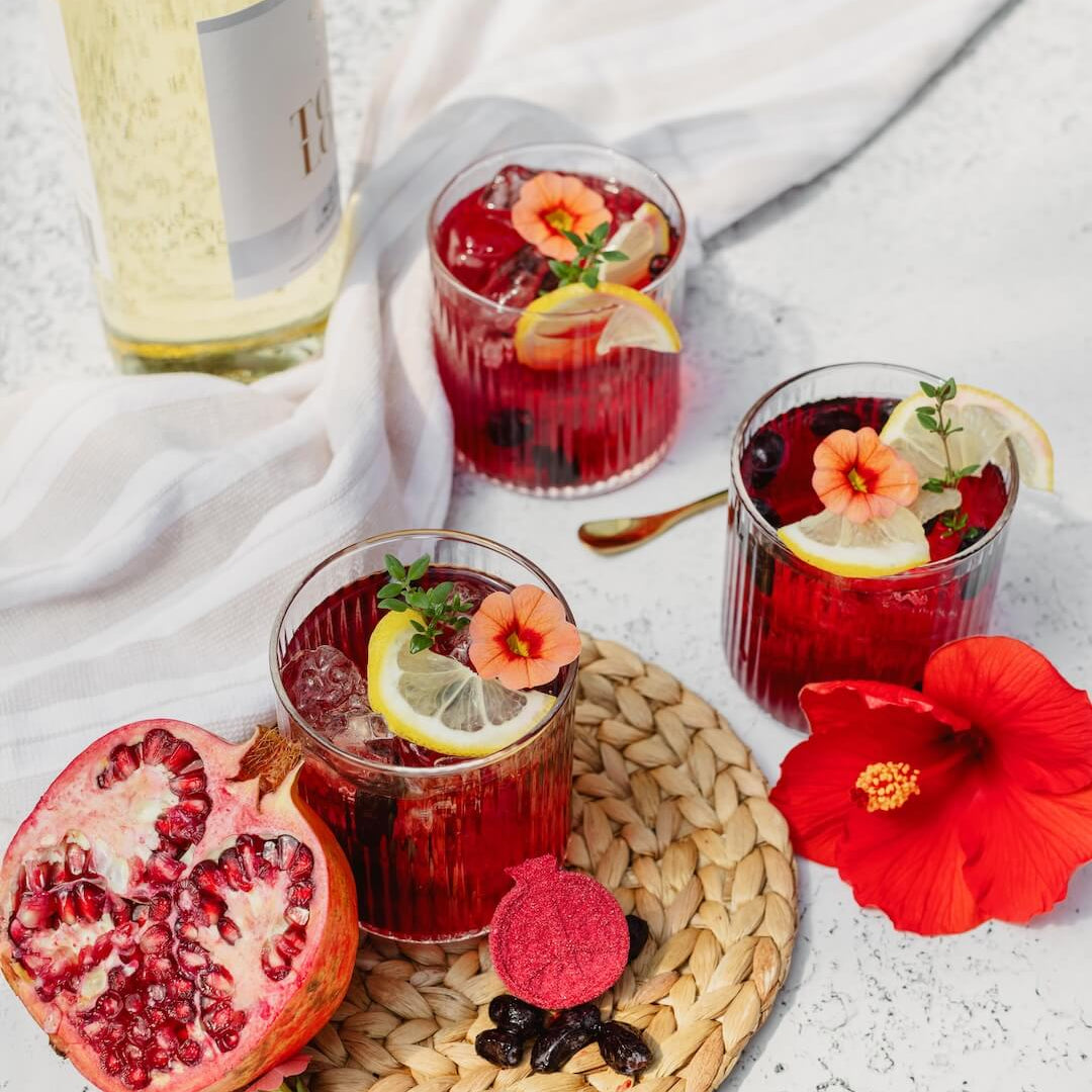 Three glasses of red cocktails with garnishes on a textured surface, accompanied by a pomegranate and a red flower.