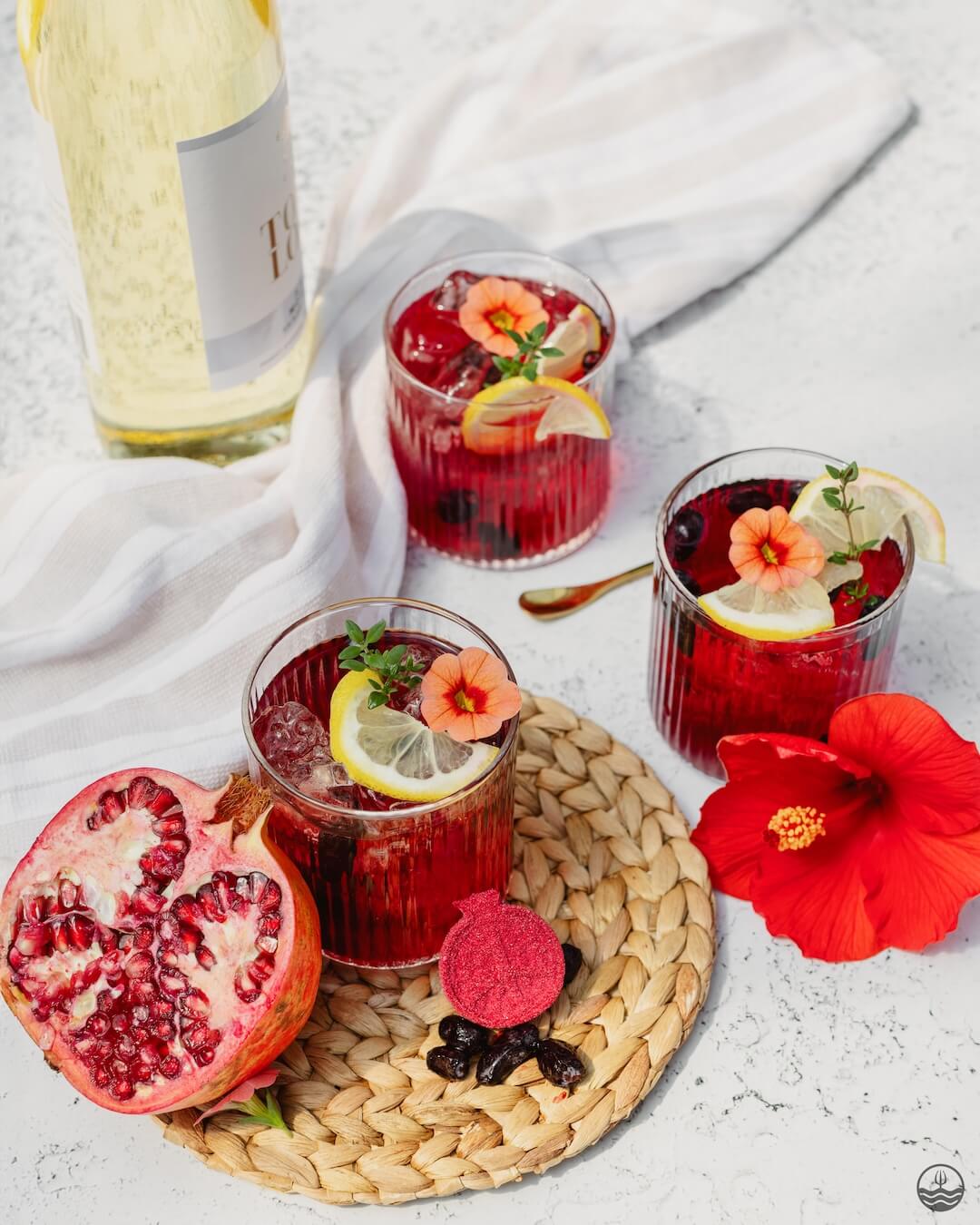 Three glasses of red cocktails with garnishes on a textured surface, accompanied by a pomegranate and a red flower.