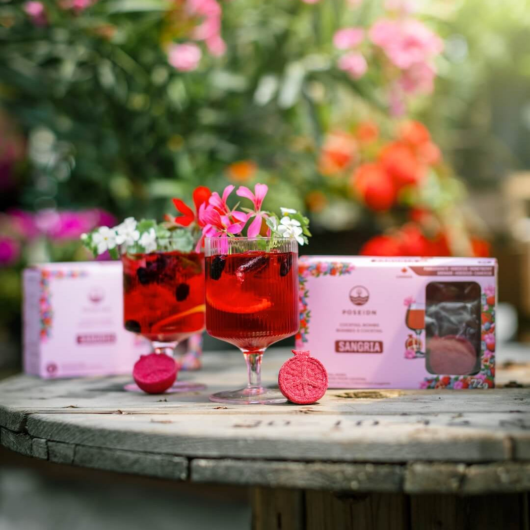 Two glasses of red cocktails with floral garnishes on a wooden table, surrounded by pink flowers and a box of Pernod Absinthe Sangria.
