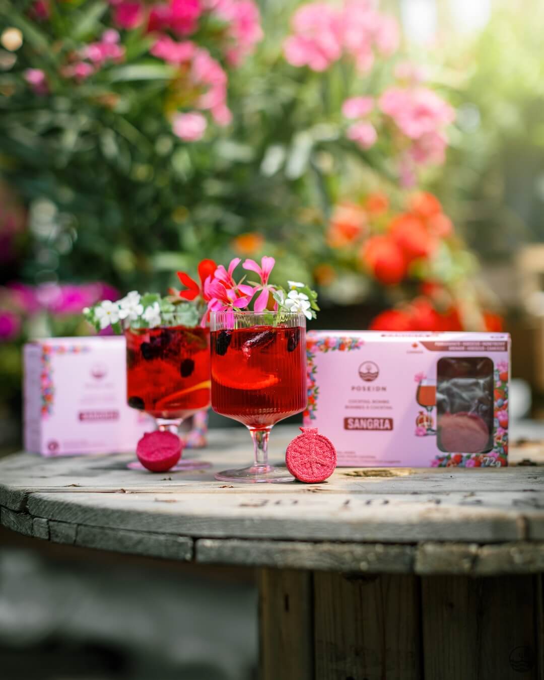 Two glasses of red cocktails with floral garnishes on a wooden table, surrounded by pink flowers and a box of Pernod Absinthe Sangria.