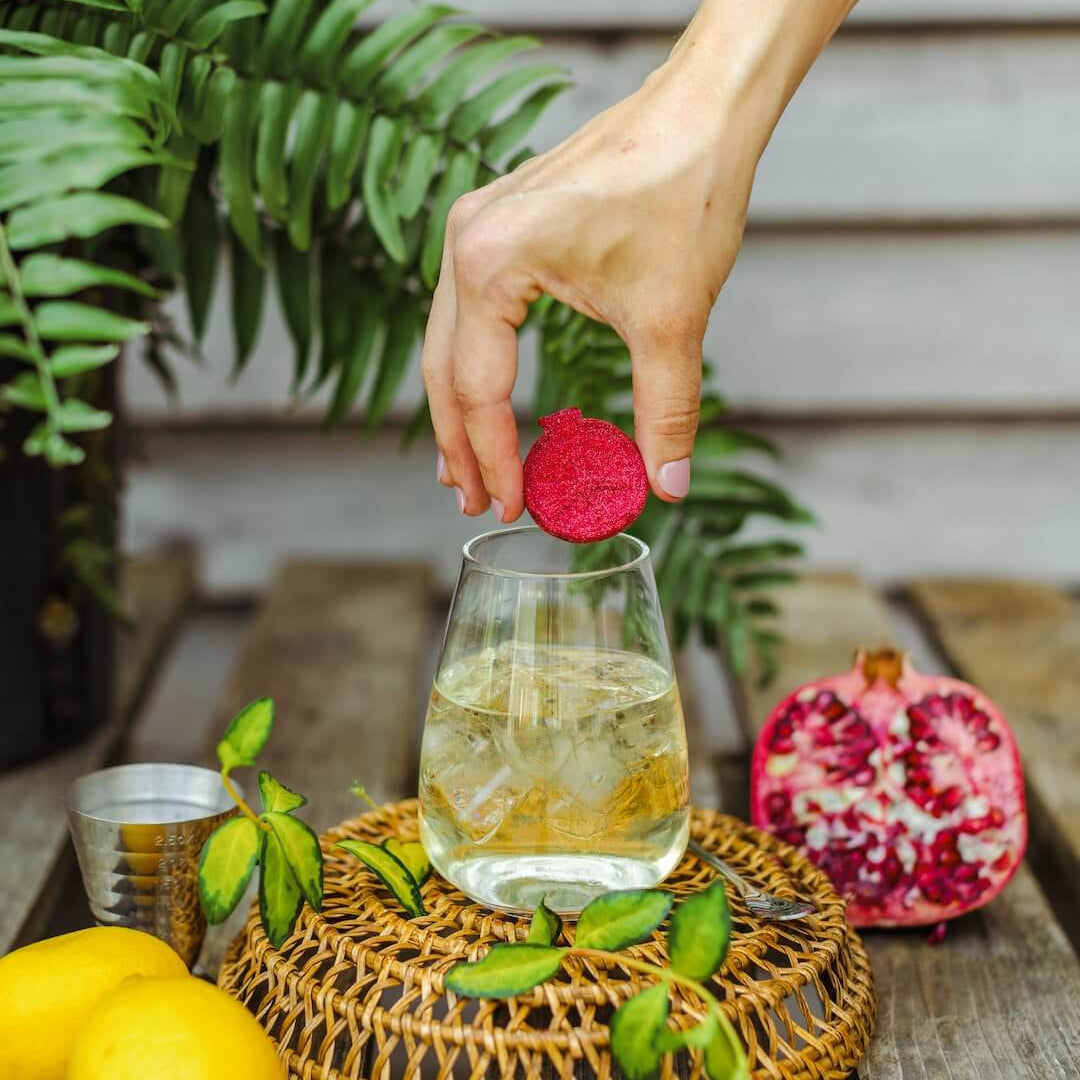 Hand placing a red tea bag into a glass of iced tea on a wooden table with lemons and a pomegranate.