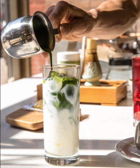 Person pouring a drink into a glass with ice and green herbs on a kitchen counter.