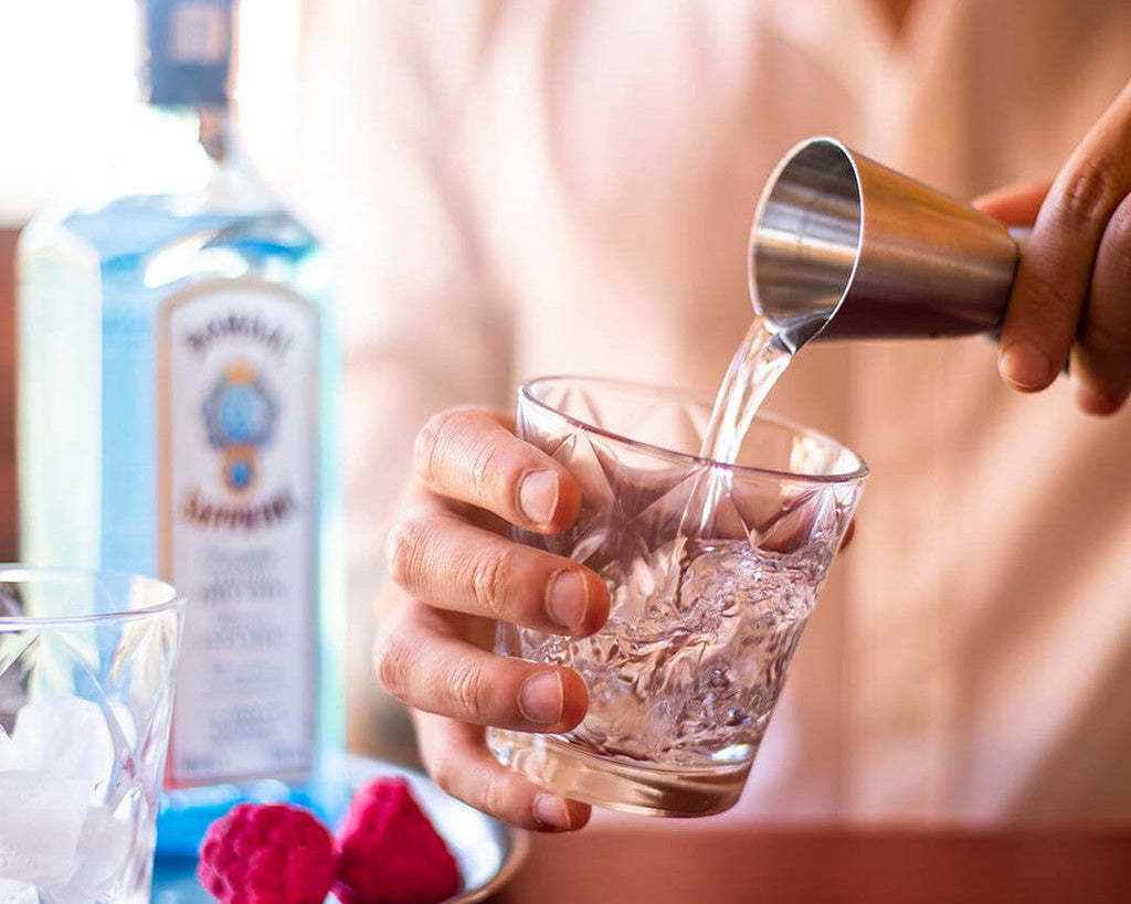 Person pouring a drink from a jigger into a glass with ice, with a bottle of vodka in the background.