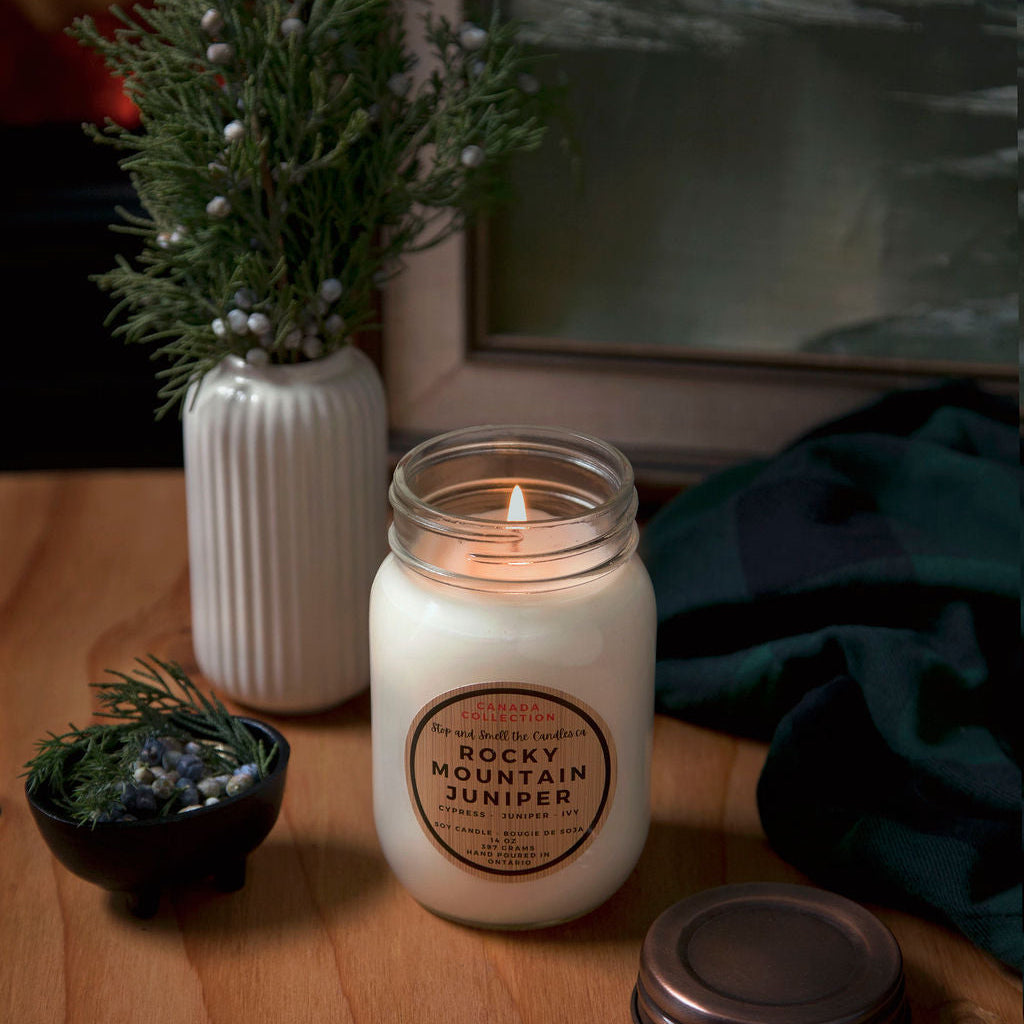 Candle with a label on a wooden surface next to a vase with greenery and a framed picture.