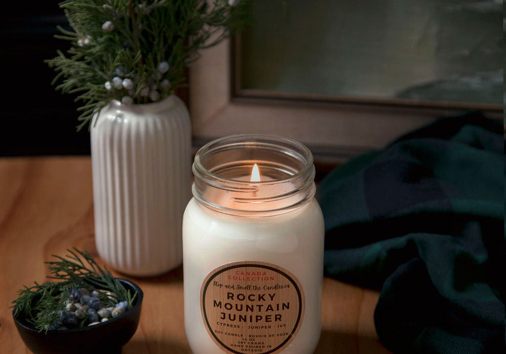 Candle with a label on a wooden surface next to a vase with greenery and a framed picture.