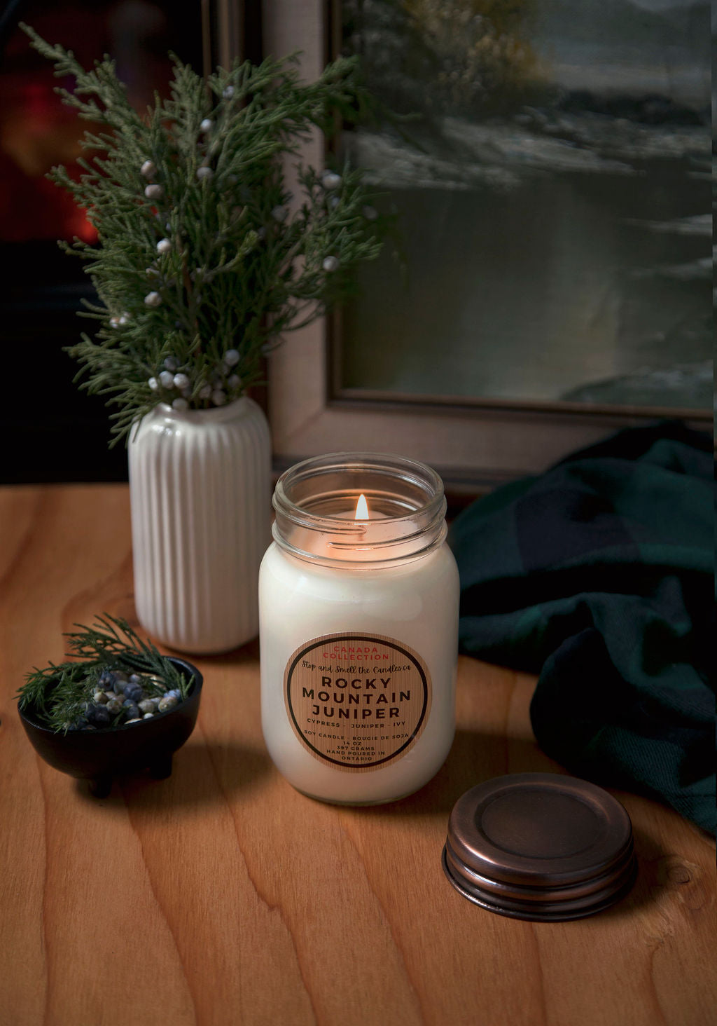 Candle with a label on a wooden surface next to a vase with greenery and a framed picture.