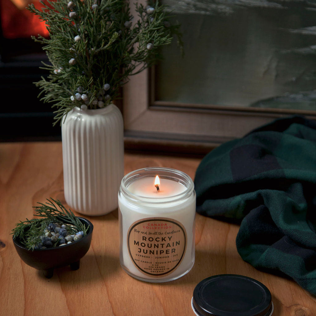 Stopandsmellthecandles Rocky Mountain Juniper soy candle – 8 oz clear glass jar, lit beside juniper branches