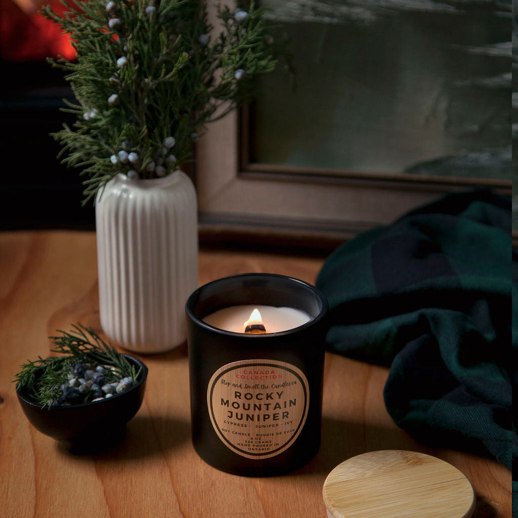 Candle with decorative packaging on a wooden surface, surrounded by plants and a towel.