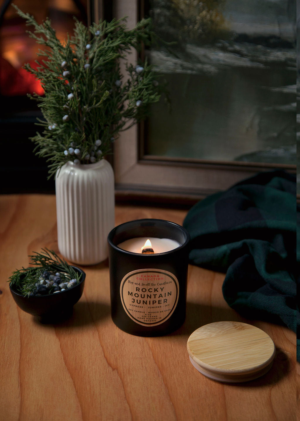 Candle with decorative packaging on a wooden surface, surrounded by plants and a towel.