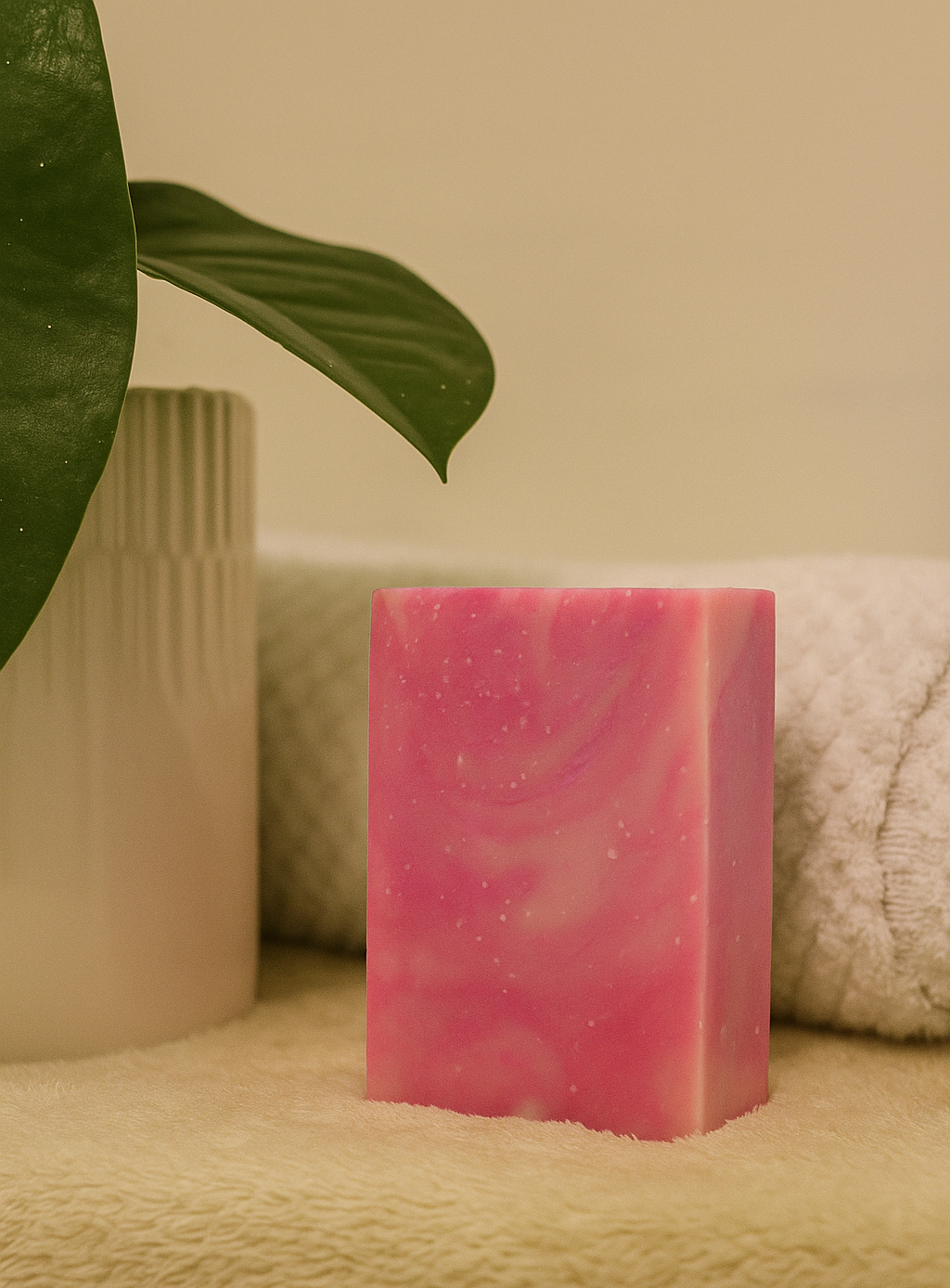 Pink soap bar on a textured surface with a plant leaf and cylindrical container in the background.