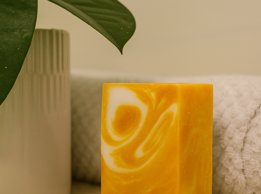 Yellow soap bar with white swirls on a beige surface, surrounded by green leaves and a textured white container.