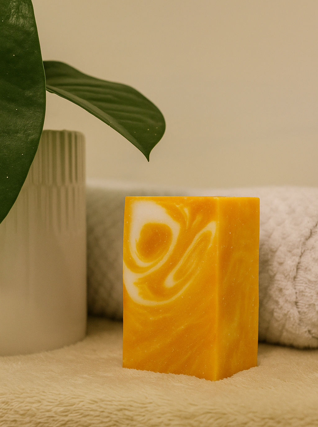 Yellow soap bar with white swirls on a beige surface, surrounded by green leaves and a textured white container.