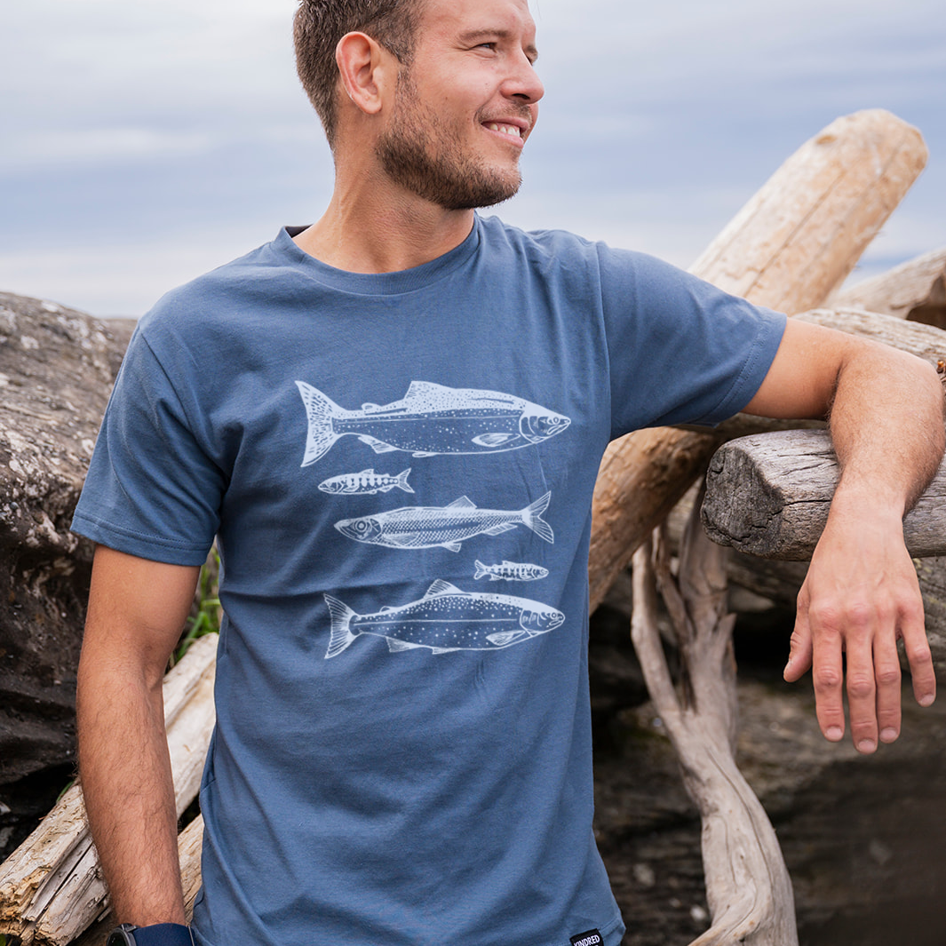 Man wearing a blue t-shirt with fish graphics, standing outdoors near wooden logs.