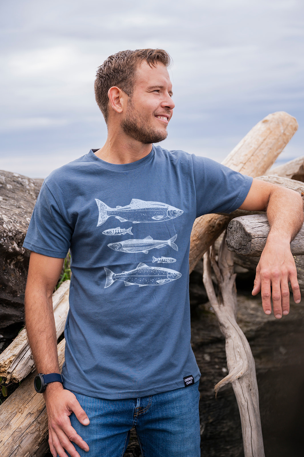 Man wearing a blue t-shirt with fish graphics, standing outdoors near wooden logs.