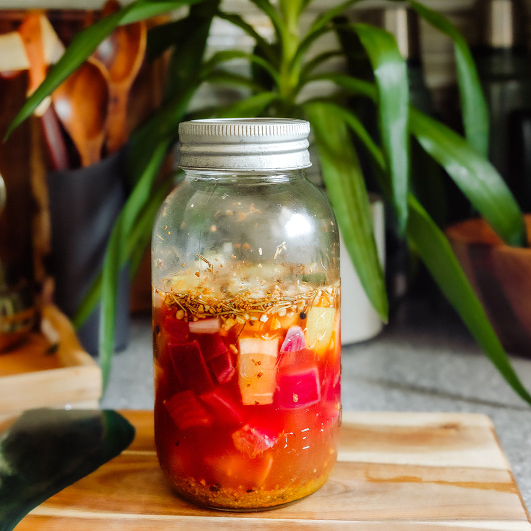Glass jar with a metal lid containing a colorful salad on a wooden cutting board with plants in the background.
