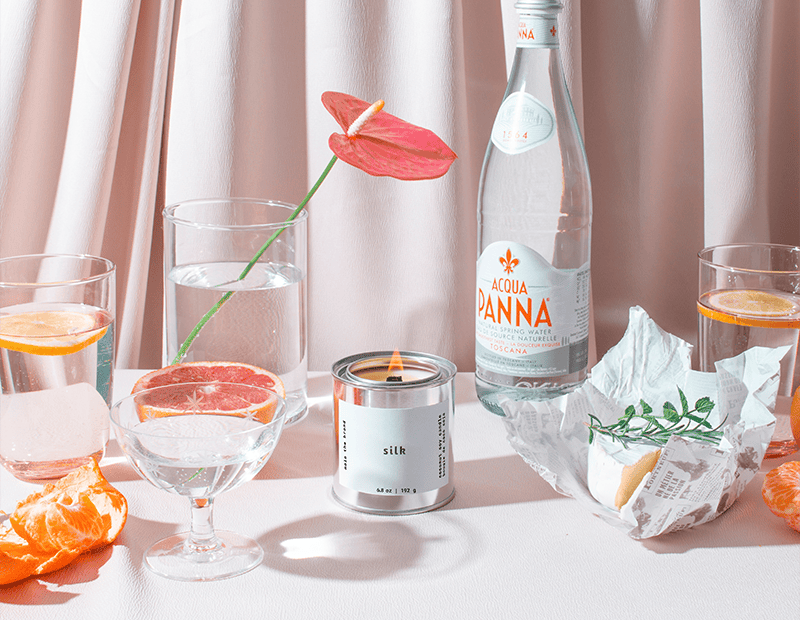 Table setting with a bottle of Voda Panina, glasses with orange slices, a candle, and a flower against a light curtain background.