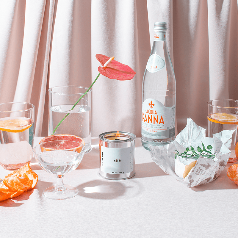 Table setting with a bottle of Voda Panina, glasses with orange slices, a candle, and a flower against a light curtain background.