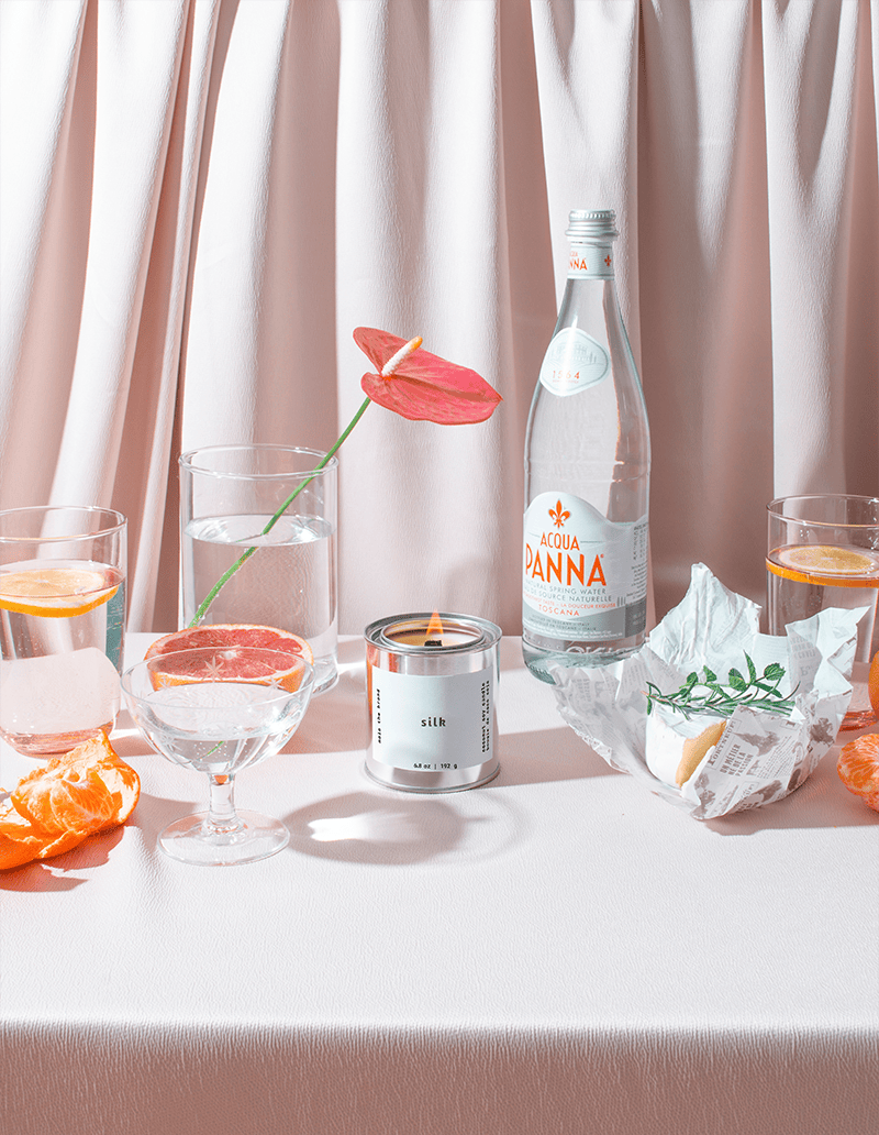 Table setting with a bottle of Voda Panina, glasses with orange slices, a candle, and a flower against a light curtain background.