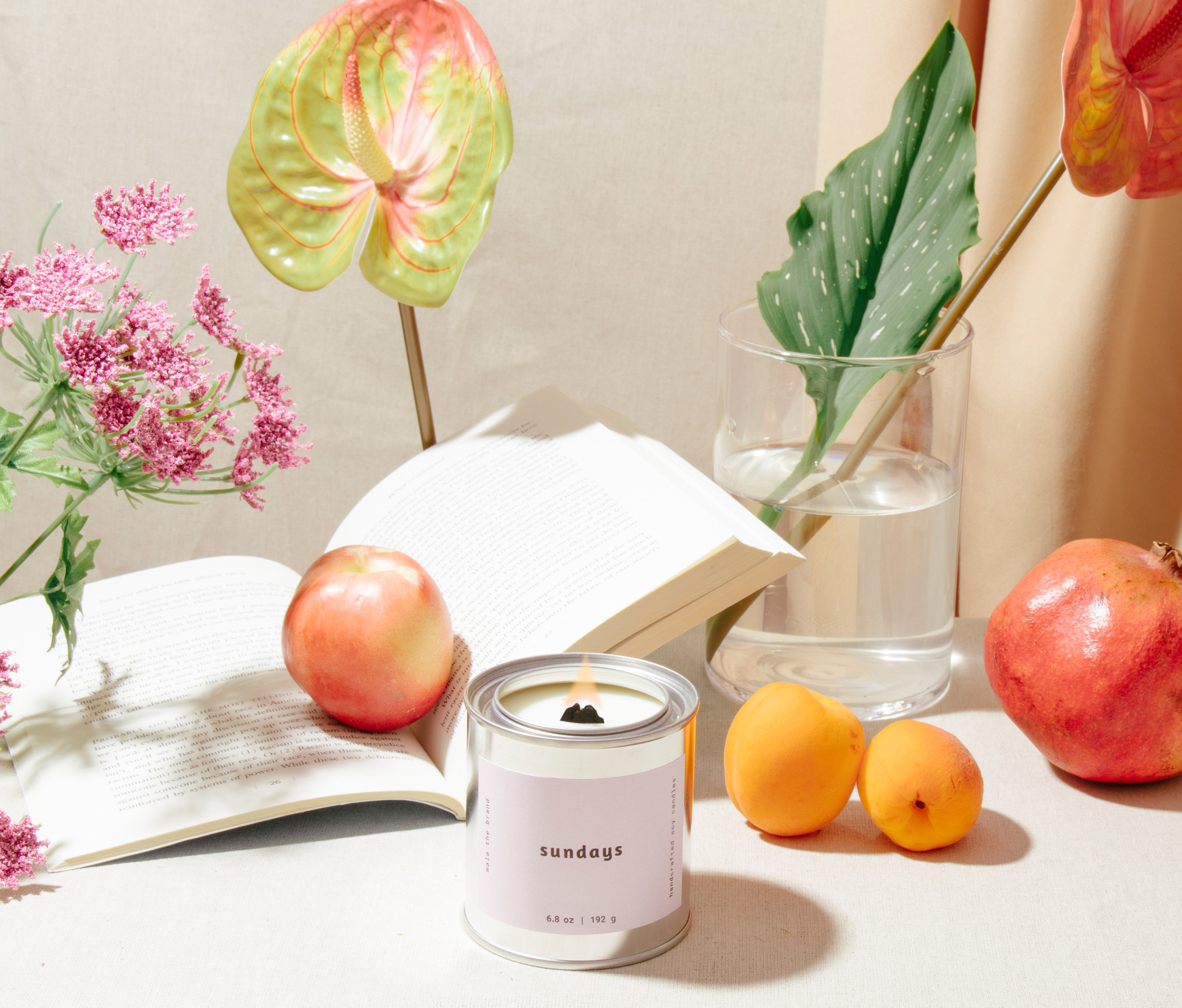 Candle with a 'sundays' label on a table with apples, oranges, and flowers.