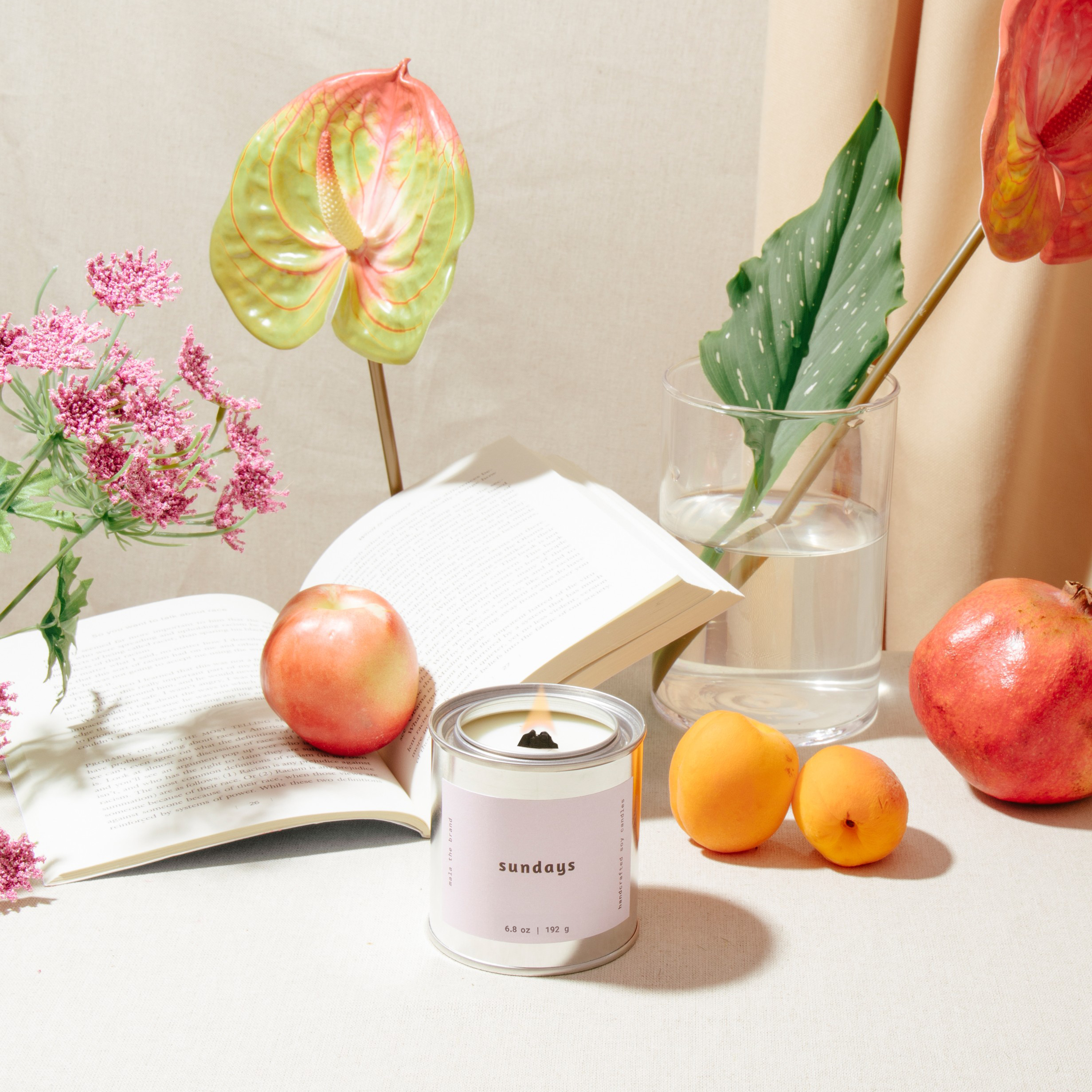 Candle with a 'sundays' label on a table with apples, oranges, and flowers.