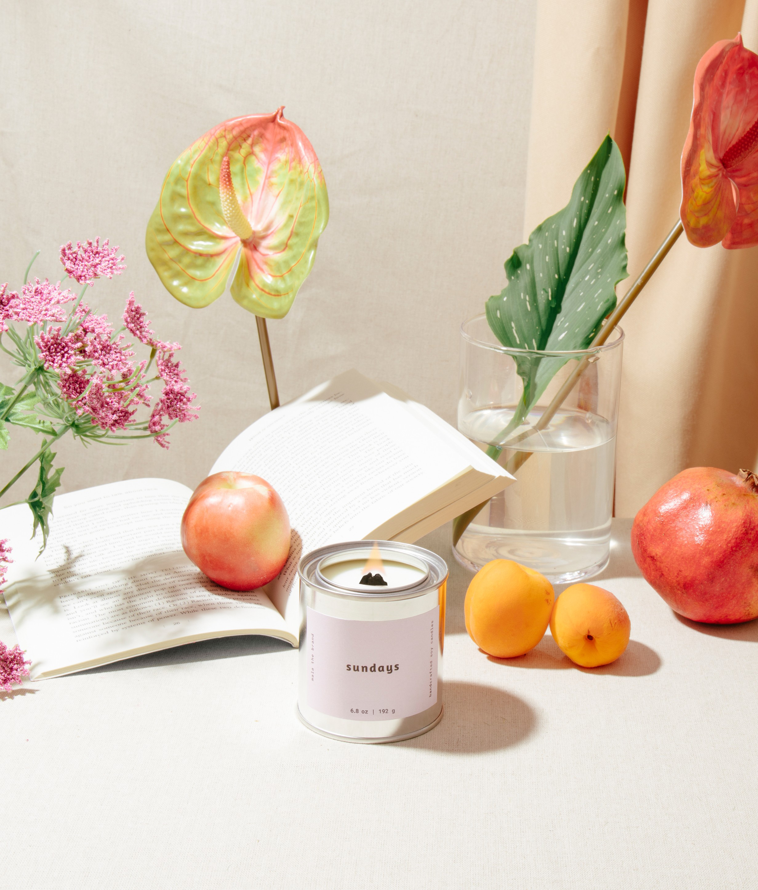 Candle with a 'sundays' label on a table with apples, oranges, and flowers.