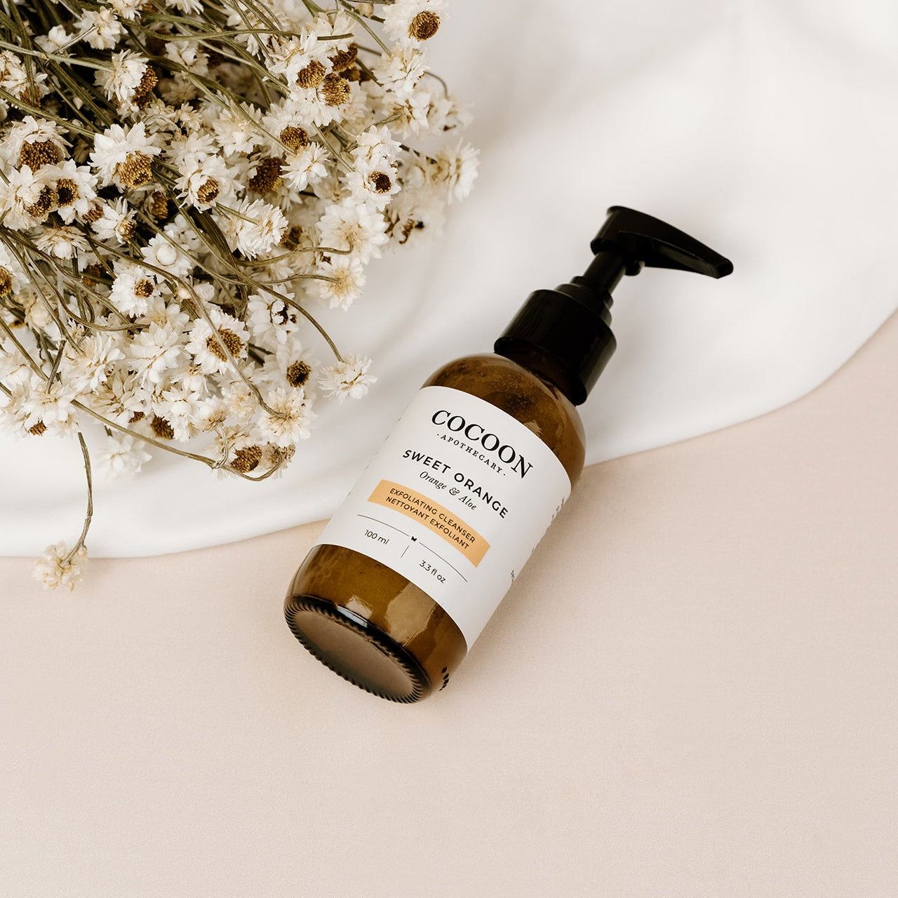 Cocoon Sweet Orange bottle on a beige surface with white flowers in the background