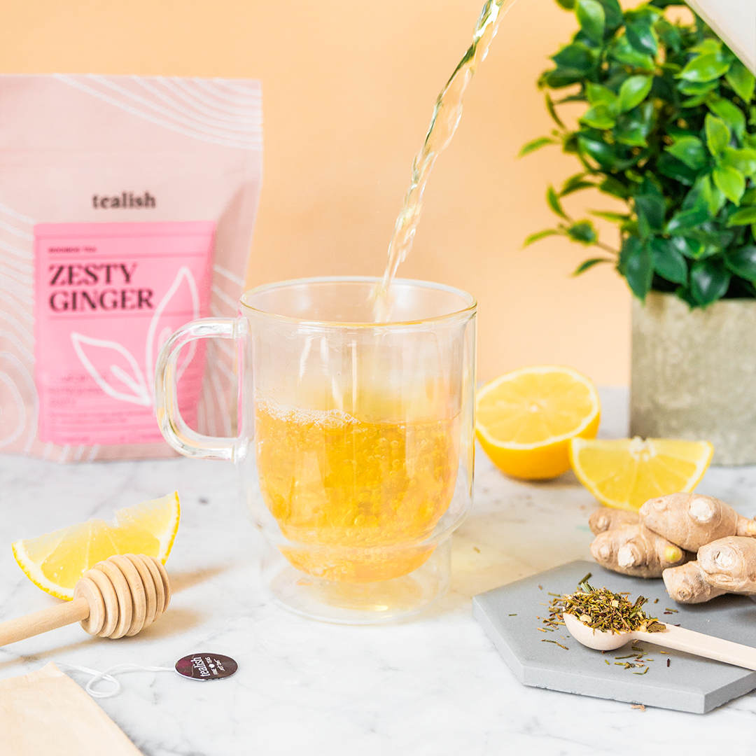 Tea being poured into a glass mug with lemon slices, ginger, and honey on a table.