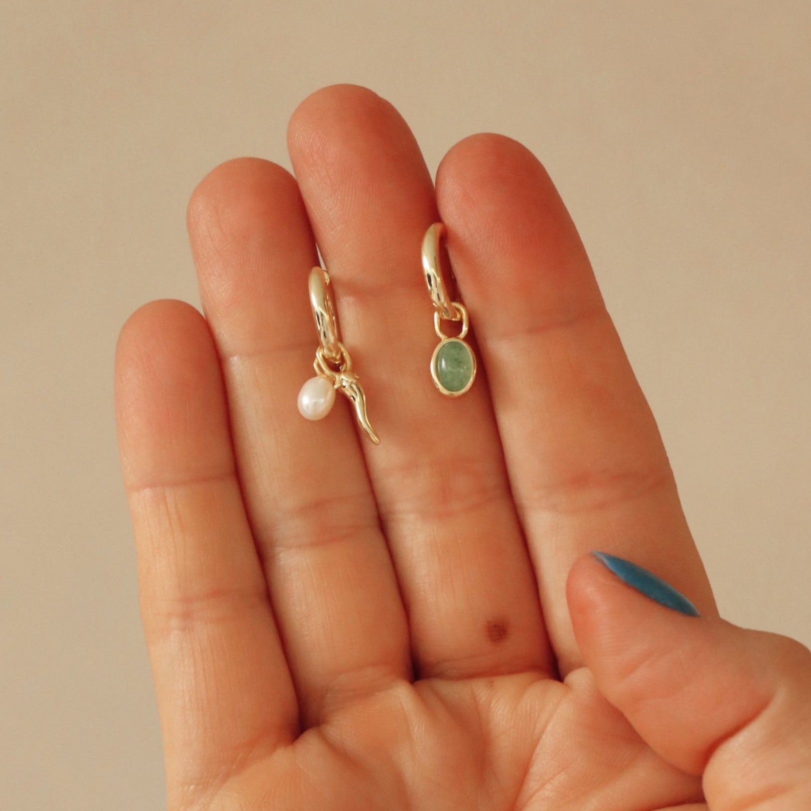 Two gold earrings with a pearl and green stone held between fingers against a beige background