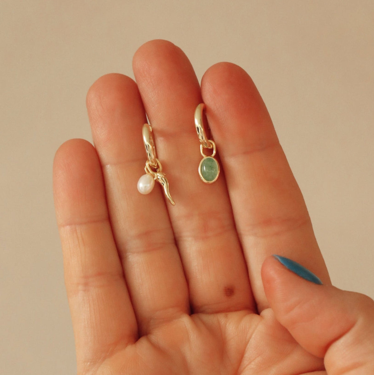 Two gold earrings with a pearl and green stone held between fingers against a beige background
