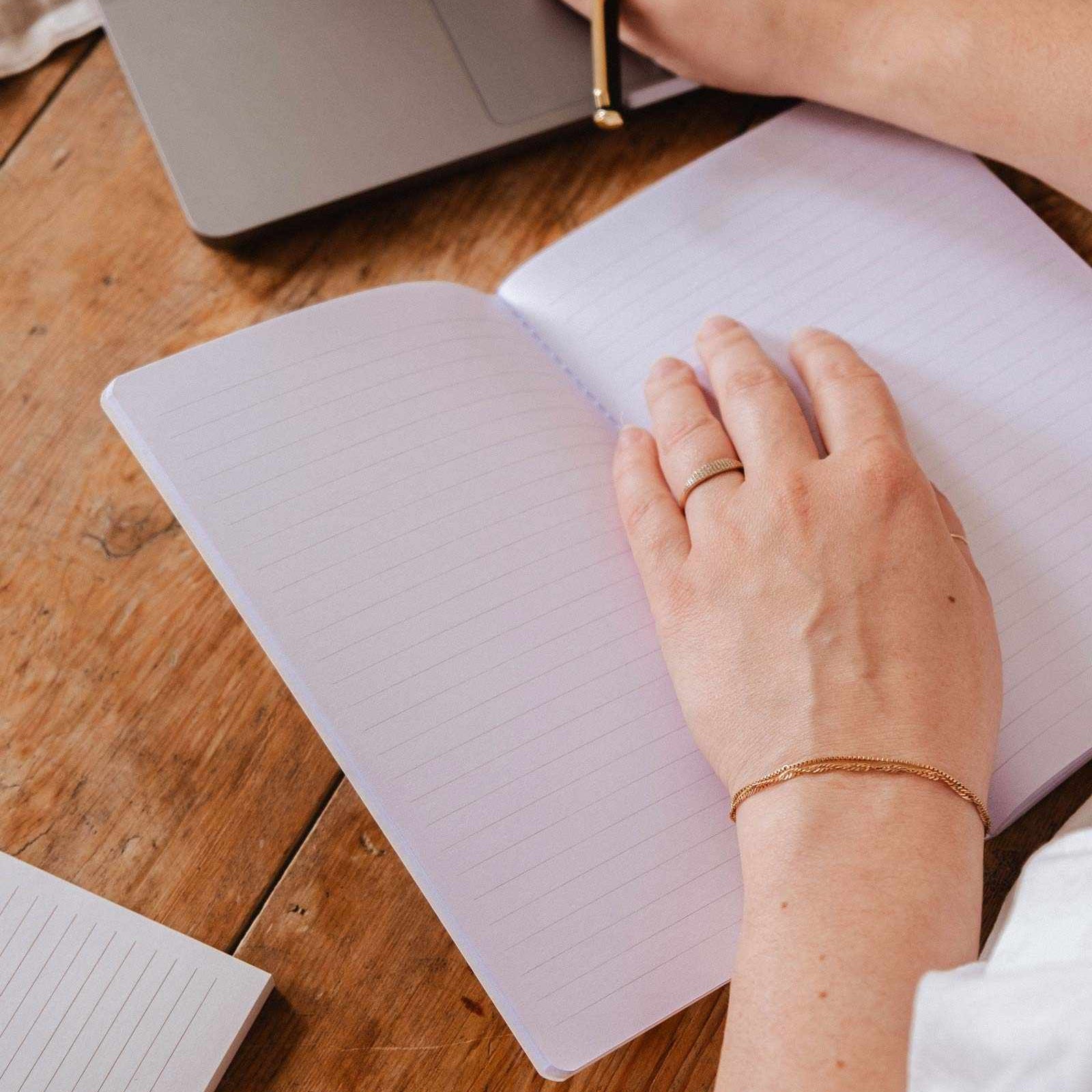 Person writing in a notebook with a laptop on a wooden desk mimi-and-august