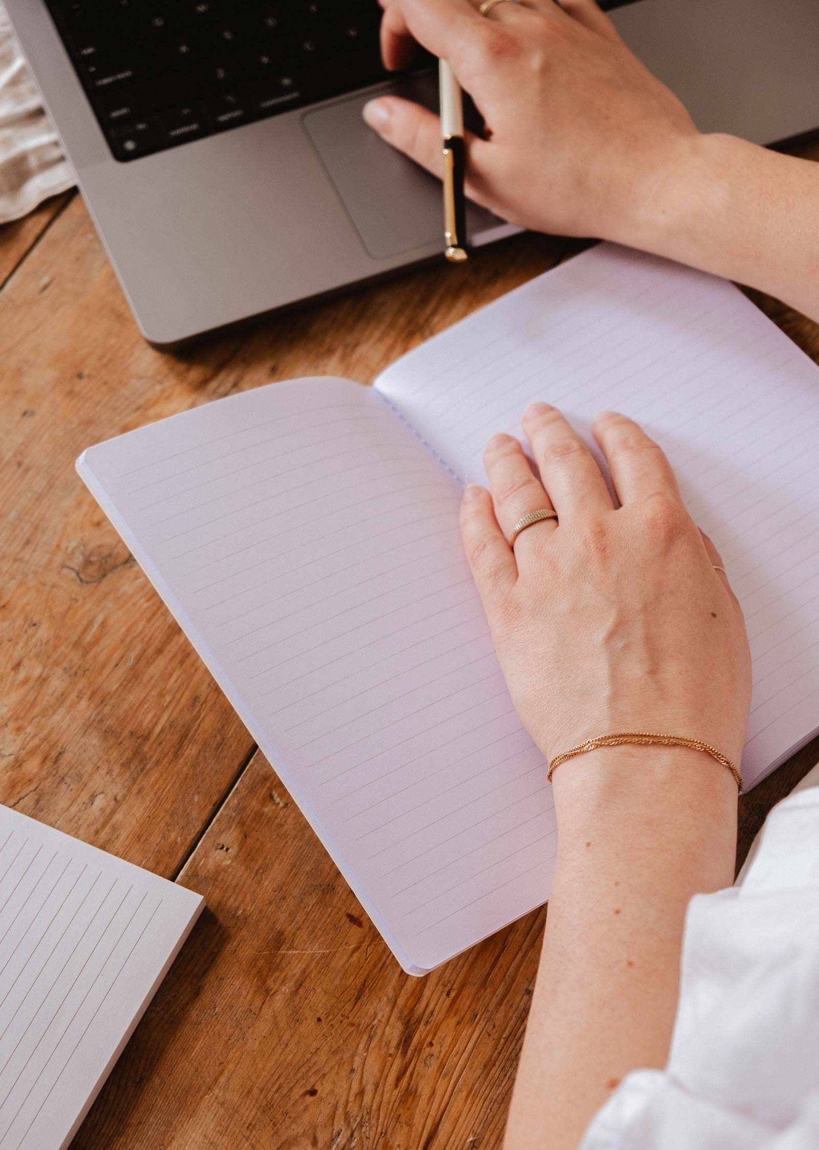 Person writing in a notebook with a laptop on a wooden desk mimi-and-august