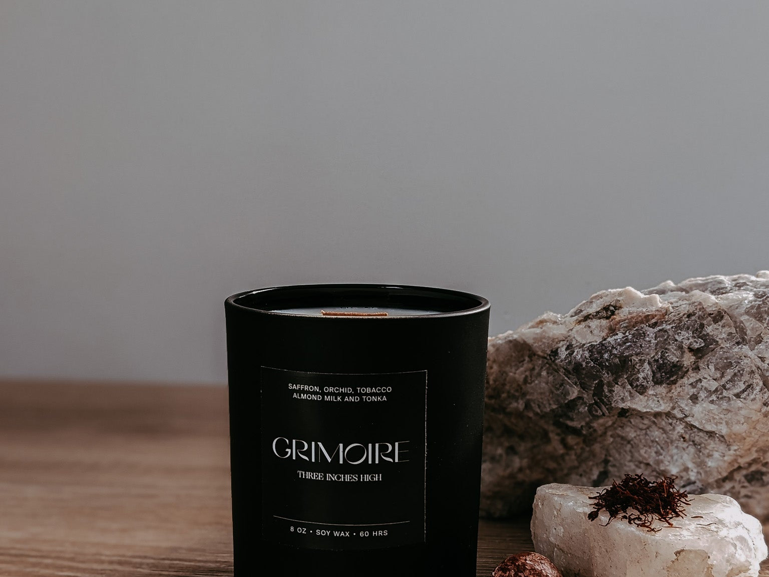 Black candle labeled 'GRIMOIRE' on a wooden surface with rocks and a neutral background