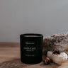 Black candle labeled 'GRIMOIRE' on a wooden surface with rocks and a neutral background
