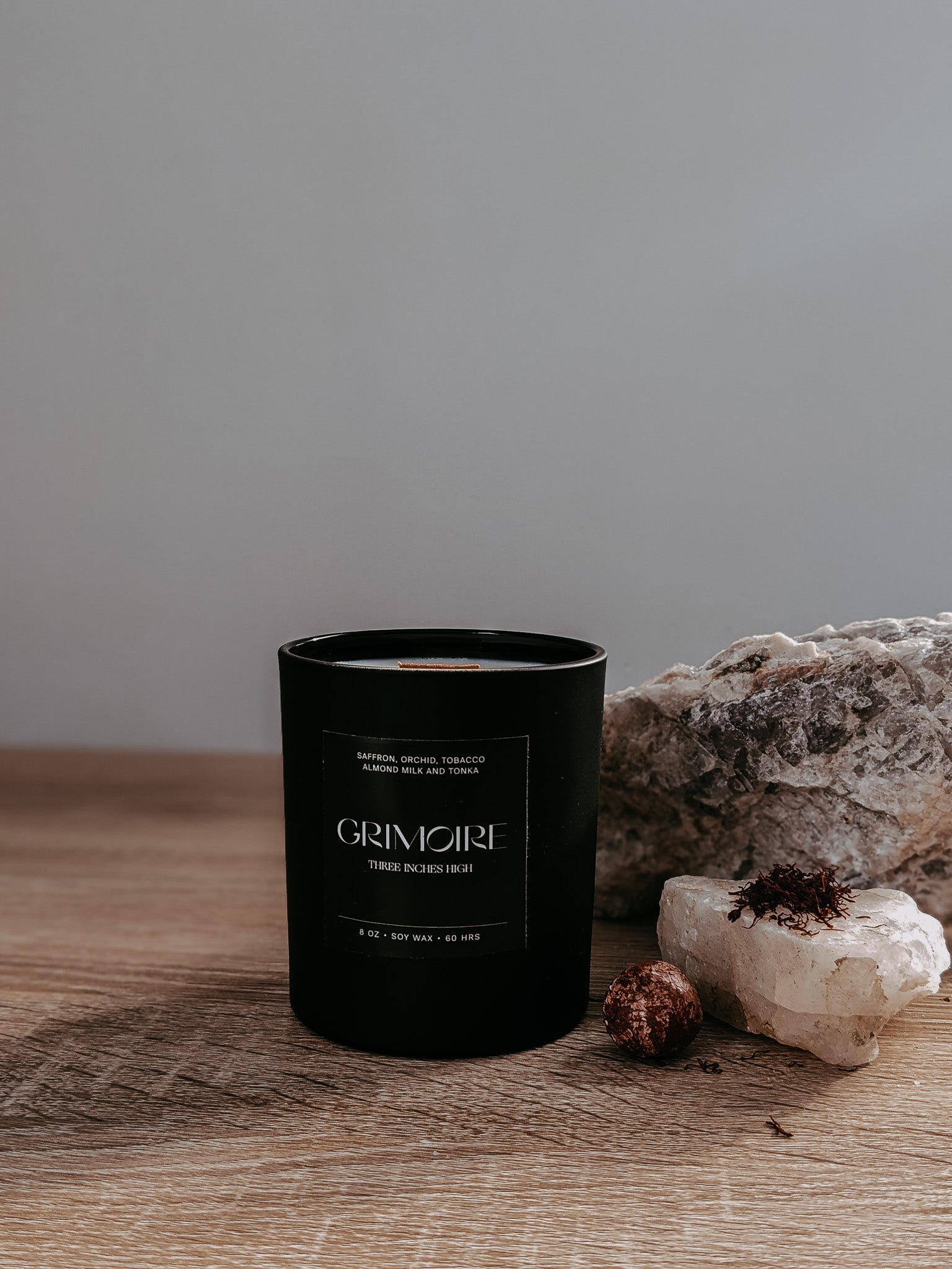 Black candle labeled 'GRIMOIRE' on a wooden surface with rocks and a neutral background