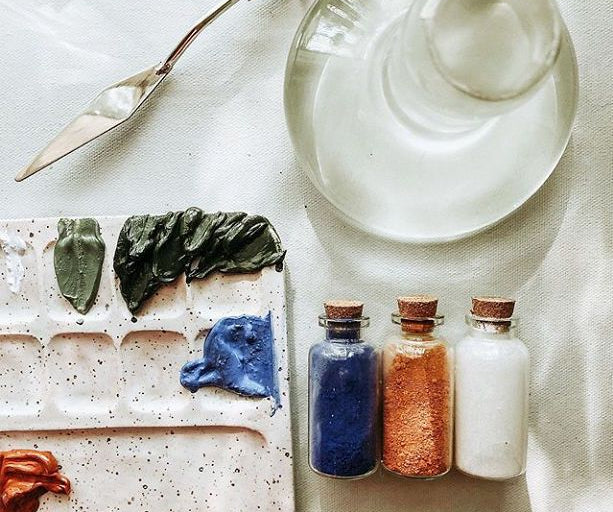 Art supplies including a palette, Clear Glass Paint Muller, paintbrush, and small bottles of paint on a textured surface.