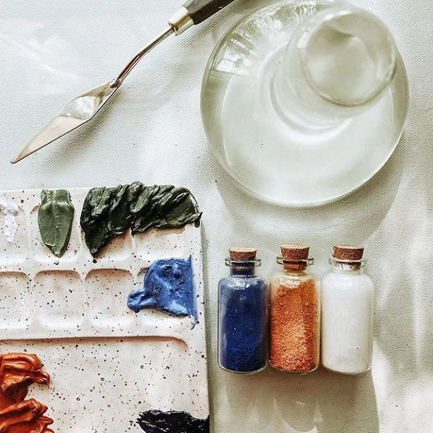 Art supplies including a palette, Clear Glass Paint Muller, paintbrush, and small bottles of paint on a textured surface.