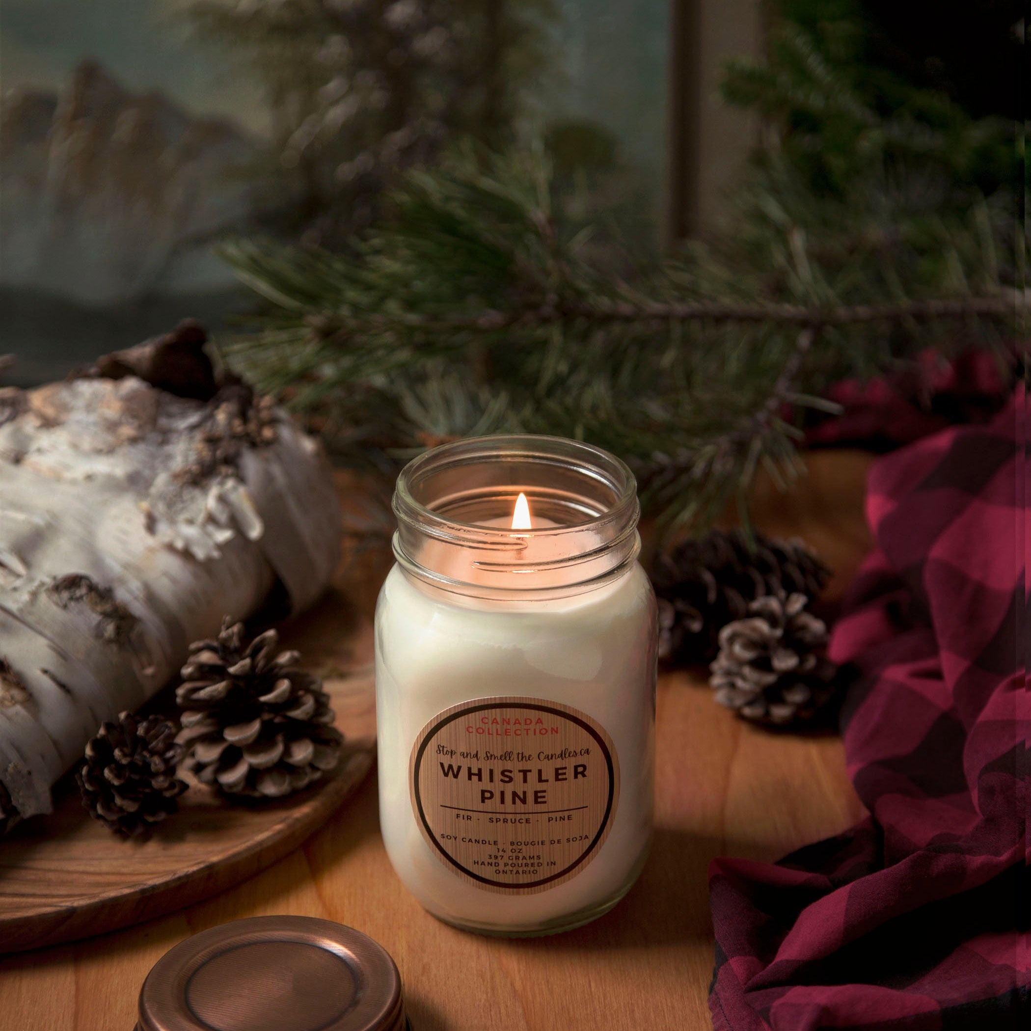Candle in a mason jar labeled 'Whistler Pine' on a wooden surface with decorative elements.
