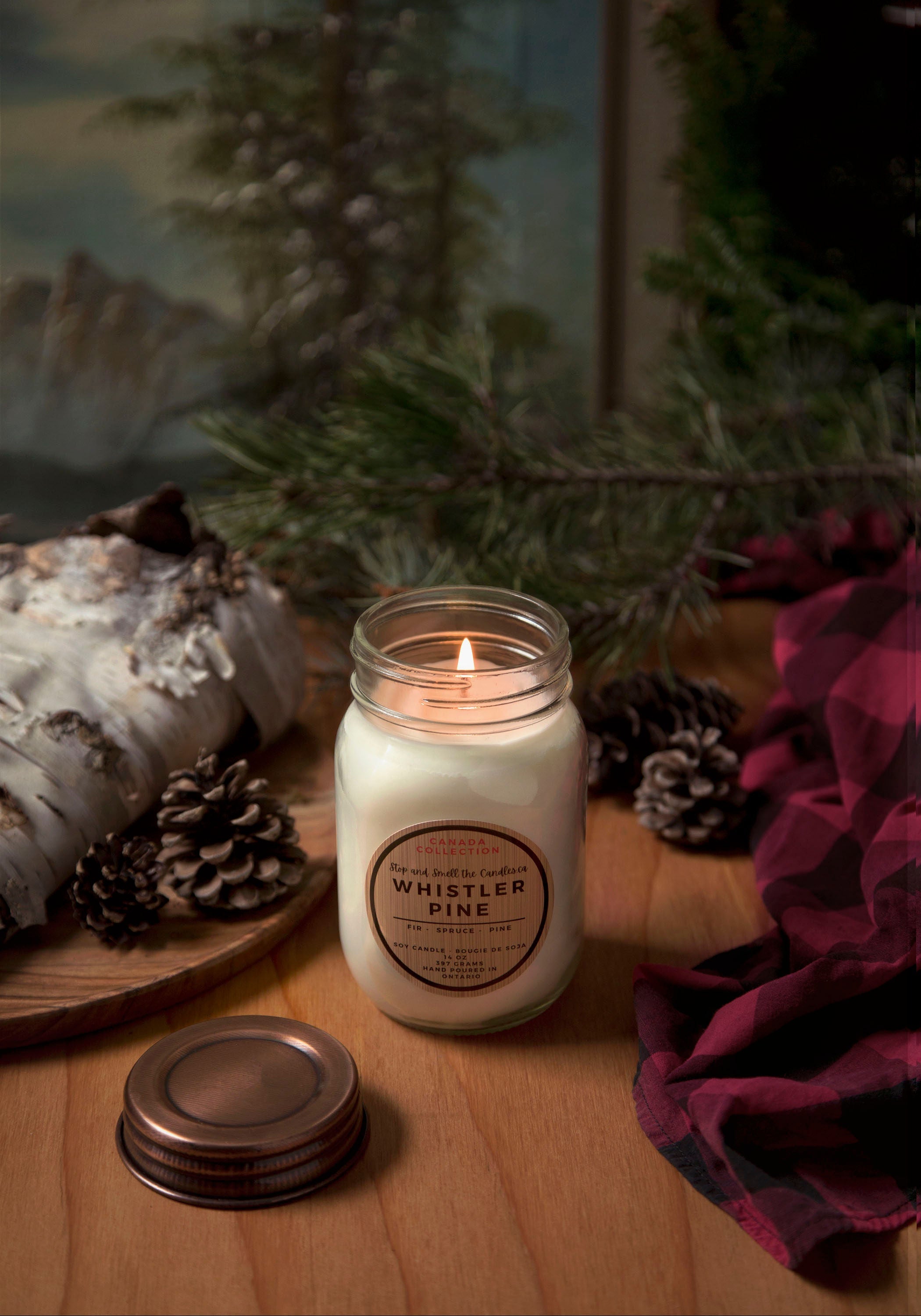 Candle in a mason jar labeled 'Whistler Pine' on a wooden surface with decorative elements.