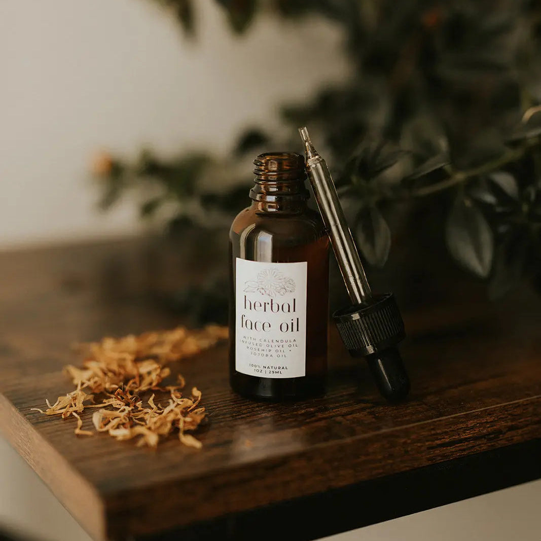 Herbal face oil bottle with dropper on a wooden surface with dried herbs and greenery in the background.