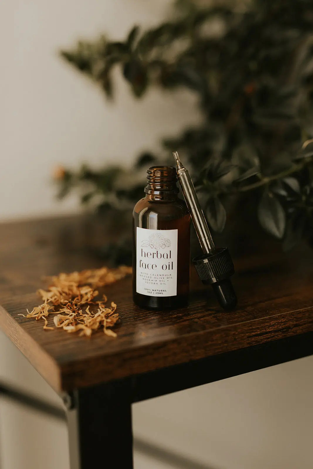 Herbal face oil bottle with dropper on a wooden surface with dried herbs and greenery in the background.