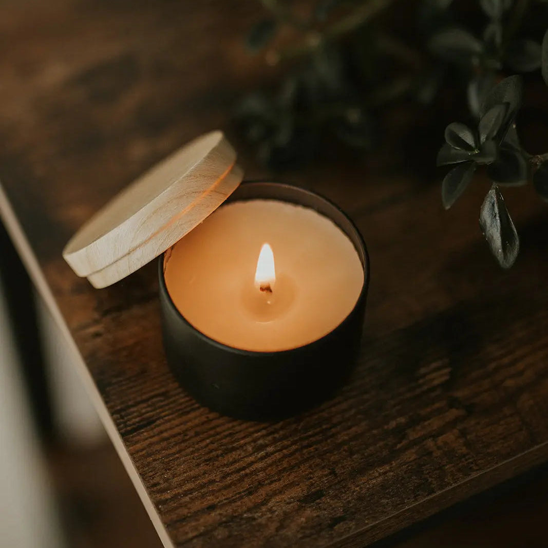 Candle in a black holder with a wooden lid on a wooden surface, surrounded by greenery.