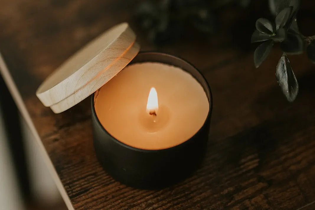 Candle in a black holder with a wooden lid on a wooden surface, surrounded by greenery.