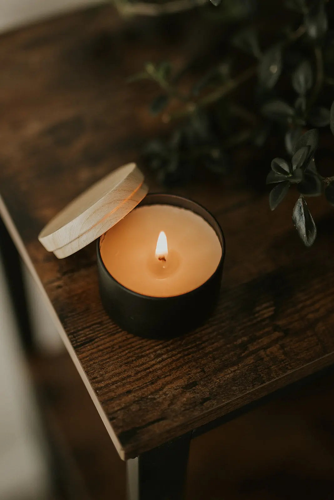 Candle in a black holder with a wooden lid on a wooden surface, surrounded by greenery.