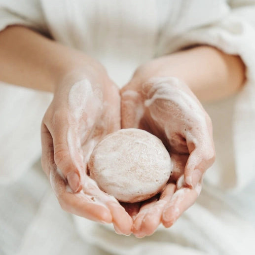 Person holding a round, beige bath bomb in their hands against a neutral background
